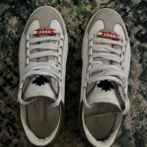 Vita sneakers från Dsquared2 - Säljer ett par vita sneakers från Dsquared2 med grå detaljer och röd Dsquared2-logga på plösen. Skorna har snörning, rund tå och platt sula. Perfekta för dig som gillar stilrena och sportiga skor.