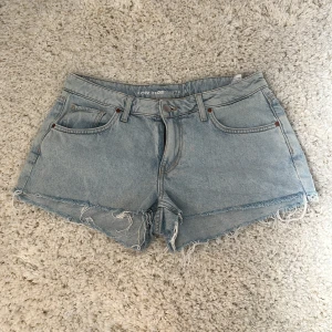 Ljusblå jeansshorts från Bikbok - Snygga ljusblå jeansshorts från Bikbok med råa, fransiga kanter och klassisk femficksdesign. Shortsen har låg midja och är tillverkade i slitstarkt denim. Liten, nästan omärkbar fläck framsida, se bild. Perfekta för sommardagar.
