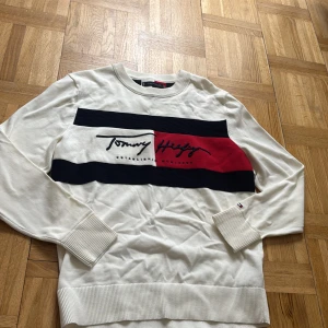 Vit stickad tröja Tommy Hilfiger S - Snygg vit stickad tröja från Tommy Hilfiger i storlek S. Tröjan har långa ärmar, breda ribbade muddar och ett stort broderat logotyptryck i svart, rött och marinblått framtill. Perfekt för dig som gillar klassisk och stilren design.