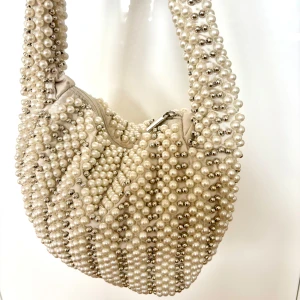 Axelväska i pärlor  - Superfin axelväska täckt av vita och silvriga pärlor. Använd bara ett fåtal gånger. Länk till väskan: https://www.na-kd.com/sv/produkter/beaded-rounded-bag-whitesilver-1100-009484-7794?_kx=MB0T9iotTF2CKZ5lJyAFnQ2YDuvJHCXfu-yFx8uG1Xw.PF6Tdw