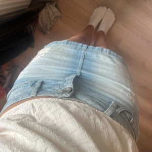 Ljusblå jeanskjol med slitningar - Säljer en ljusblå jeanskjol med snygga slitningar och klassiska fickor. Kjolen är kort och har en normal passform med knapp och dragkedja framtill. Perfekt för dig som gillar en chill och trendig stil till sommaren.