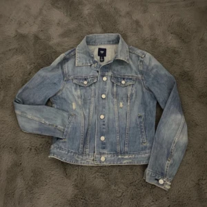 Ljusblå jeansjacka från GAP - Klassisk ljusblå jeansjacka från GAP med silvriga knappar framtill och två bröstfickor. Jackan har en rak passform och är tillverkad i denim. Perfekt att slänga över en hoodie eller t-shirt för en avslappnad look.