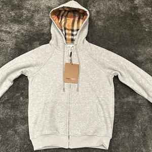 Grå Burberry kofta - Säljer en grå hoodie från Burberry med dragkedja och justerbar huva. Insidan av huvan har det ikoniska Burberry-rutiga fodret i beige, svart och rött. Tröjan är i mjuk bomull och har ribbade muddar vid ärmslut och nederkant. Den är liten i storleken och passar någon med storlek S/XS