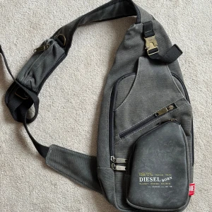 Vintage Diesel axelväska - Hej hej! Säljer min gråa dieselväska. Har inte använts särkilt mycket så den är i nästan helt perfekt skick. Den har 5 olika fack o fickor där man får plats med allt vardagligt o lite till. Storleksmässigt är den  43cm lång o 21cm bred. All hardware, som dragkedjor o så, är i mässing. 