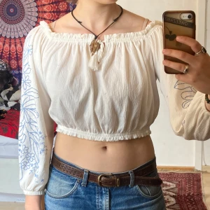 Vit croppad blus från H&M, M - Superfin sommartröja! Croppad och offsholder med långa ärmar. Blusen är vit och har blå broderade blommor på ärmarna och resår vid midjan och upptill. Har inte använts så mycket, inga defekter.