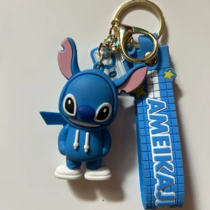Blå Stitch nyckelring med AMEIKAJI band - Supergullig blå Stitch-figur i mjuk syntet med vita sneakers och hoodie, fäst på ett blått band med texten AMEIKAJI och stjärndetaljer. Nyckelringen har även en liten kamera-charm och guldfärgad ring. Perfekt accessoar till väskan eller nycklarna!