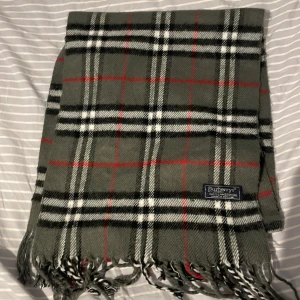 Rutig grå halsduk från Burberrys - Klassisk rutig halsduk från Burberrys i 100% lammull. Färgerna är grå, svart, vit och röd med fransar i ändarna. Perfekt accessoar för att lyfta din vinterstil med ikoniskt brittiskt mönster.