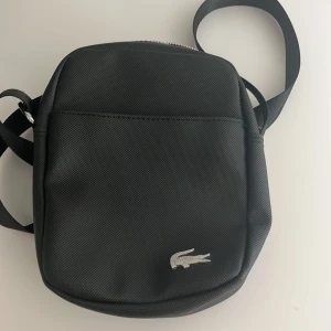 Svart axelväska från Lacoste - Snygg svart axelväska från Lacoste med klassisk krokodillogga framtill. Väskan har justerbar axelrem och dragkedja upptill. Perfekt storlek för det viktigaste och enkel att bära med sig.