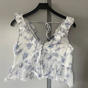Vit blommig topp med volang - Superfin vit croppad linnetopp med blått blommigt mönster och volangdetaljer vid axlar och nederkant. Toppen har knytning i ryggen och en luftig känsla, perfekt för varma dagar. Mycket bra skick o endast använd en gång💖💖