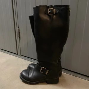 Svarta höga boots med spännen - Säljer ett par snygg svarta höga boots i skinn med grov sula och två silvriga spännen, ett vid vristen och ett upptill. Dragkedja baktill för enkel påtagning. 