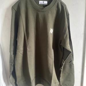 Olivgrön sweatshirt från Stone Island - Säljer en olivgrön sweatshirt från Stone Island med rund halsringning och diskret logopatch på bröstet. Tröjan är i mjuk bomull och har långa ärmar. Perfekt för en stilren och avslappnad look.