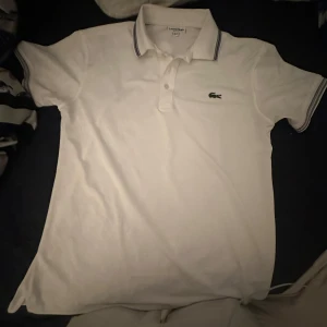 Vit piké t-shirt från Lacoste - Vit piké t-shirt från Lacoste med klassisk krage och knappar framtill. Har bara använt den 1 gång för att den är för liten. Storlek är L men går och ha Xl.