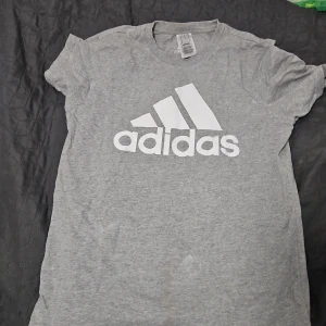 Grå t-shirt från Adidas - Säljer en grå t-shirt från Adidas med stor vit logga framtill. Klassisk rund halsringning och korta ärmar. Perfekt för en sportig och avslappnad stil.