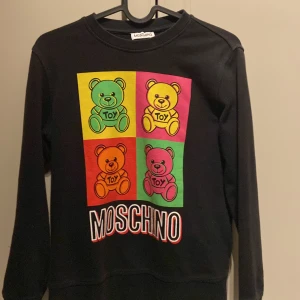 Svart Moschino tröja med nalletryck - Svart långärmad tröja från Moschino med färgglatt pop art-tryck av fyra nallar i grönt, gult, orange och rosa. Stor MOSCHINO-logga framtill. Mjuk bomullskänsla och relaxed fit, perfekt för dig som gillar statement-plagg.