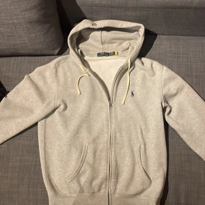 Grå hoodie från Polo Ralph Lauren - Snygg ljusgrå hoodie från Polo Ralph Lauren med dragkedja, huva med snörning och klassisk blå logga på bröstet. Tillverkad i mjuk bomullsmix och har två fickor framtill. Perfekt för en chill och stilren look.