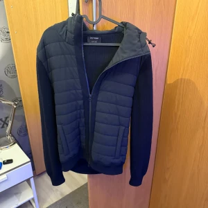 Gran Sasso marinblå pufferjacka - Marinblå pufferjacka från Gran Sasso med stickade ärmar och quiltad front. Jackan har huva med dragsko och hel dragkedja. Tillverkad i Italien i en mix av ull och syntetmaterial. Perfekt för dig som gillar stilren och sportig look. 