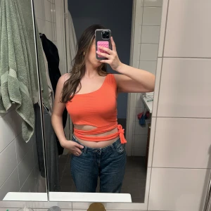 Korall färg oneshoulder croppad topp med knytband  - Säljer en orange croppad topp med oneshoulder-design och knyt i sidan. Toppen har en cut-out detalj vid midjan och är ärmlös. Perfekt för dig som gillar trendiga och unika plagg med snygg passform. Mycket bra skick, storlek S.🩷