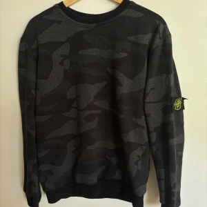 Svart camouflagemönstrad sweatshirt från Stone Island - Säljer en svart och grå camouflagemönstrad sweatshirt från Stone Island med klassisk logotyp-patch på vänster ärm. Tröjan har rund halsringning och ribbade muddar vid ärmslut och nederkant. Perfekt för en streetwear-look.