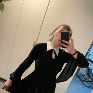 Svart långärmad klänning med vit krage från Killstar - Säljer en svart långärmad klänning från Killstar med vit spetsig krage och utsvängda ärmar. Klänningen har en liten öppning framtill under kragen och är gjord i ett sammet material.