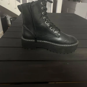 Svarta boots med snörning och dragkedja - Säljer ett par svarta boots med snörning och dragkedja på sidan. Skorna har en kraftig platt sula och rund tå. Perfekta för dig som vill ha en tuff och stilren look.