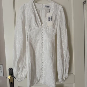 Vit långärmad klänning från Adoore - Helt ny, oanvänd med prislappen på! Supersnygg vit klänning från Adoore med långa puffärmar, v-ringning och dekorativa knappar framtill. Klänningen har ett subtilt blommigt mönster och är perfekt för sommarens alla tillfällen. Tillverkad i ett lätt och bekvämt material.