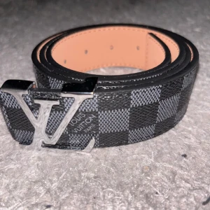 Louis Vuitton  bälte i skinn - Snyggt Louis Vuitton-bälte i svart och grått med klassiskt rutigt Damier-mönster. Bältet har en stor LV-logga i silvermetall som spänne och är gjort i exklusivt skinn. Perfekt accessoar för att lyfta din outfit med en lyxig touch. 