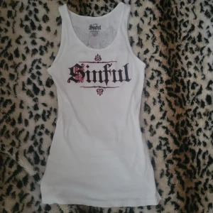 Vit linnetopp från Sinful med tryck - Vit ribbad linnetopp från Sinful med svart och rött tryck framtill där det står 'Sinful' i gotisk stil. Baksidan har ett stort tryck med svarta änglavingar och ett rött S. Linnetoppen är en L men passar mig som är en S. Skriv om du har frågor😁