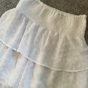 Vit kortkjol med volang och prickigt mönster - Superfin vit kortkjol med bred smock i midjan och dubbla volanger. Kjolen har ett diskret prickigt mönster i tyget och är perfekt för varma dagar. Lätt och luftig känsla.