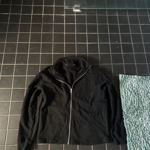 Svart jacka med dragkedja - En svart jacka med hel dragkedja framtill och hög krage. Jackan har lång ärm och en enkel, stilren design utan synliga loggor eller mönster. Passar perfekt för dig som gillar en clean look.