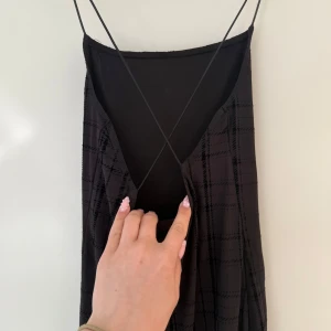 Svart klänning med tunna axelband - Säljer en svart klänning med tunna axelband och diskret rutigt mönster. Klänningen har djup v-ringning i ryggen och smal passform som följer kroppen. Perfekt för dig som gillar minimalistisk och stilren look.
