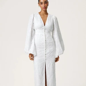 Vit långklänning med puffärmar och broderade detaljer - Adoore positano dress. Superfin vit långklänning med djup v-ringning, puffiga långärmar och knappar framtill. Klänningen har broderade blommor i tyget och markerad midja. Perfekt för sommarens speciella tillfällen! Nypris 1695kr