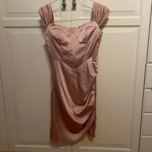 Rosa satin klänning med drapering - Säljer en rosa kort klänning i satin material med korsett byst, breda axelband och snygg drapering på sidan. Klänningen har en figurnära passform och en liten slits för extra detalj i strl S