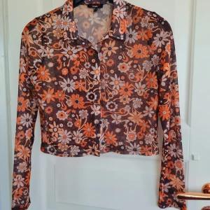 Blommig meshskjorta från Jennyfer - Säljer en transparent, blommig skjorta från Jennyfer i meshmaterial. Skjortan har långa ärmar, krage och knäppning framtill. Färgerna går i orange, svart och vitt med ett tydligt blommönster. Perfekt för dig som gillar färg och mönster!