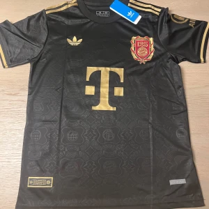 Svart Bayern München fotbollströja Musiala 42 - Snygg svart Bayern München fotbollströja med guldiga detaljer, Adidas-logga och klubbmärke på bröstet. Nummer 42 och namnet Musiala på ryggen. Korta ärmar och klassisk passform. Perfekt för fans!