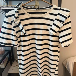 Randig ribbad t-shirt med volang från Zara - Kortärmad ribbad t-shirt från Zara i vitt och svart med ränder. T-shirten har volangdetaljer på axlarna och en rund halsringning. Perfekt för en trendig och avslappnad look. Passar mig utmärkt som är en storlek S!