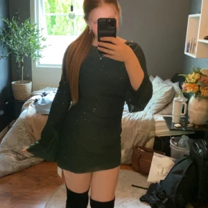 Mörkgrön glittrig klänning med utsvängda ärmar - Snygg mörkgrön klänning med glittrigt tyg och långa utsvängda ärmar. Kort modell som sitter tight och har en enkel rund halsringning. Perfekt för dig som vill sticka ut lite extra.
