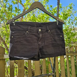Svarta jeansshorts - Säljer ett par svarta jeansshorts med klassisk femficksdesign och knappgylf. Shortsen har låg midja och är perfekta för varma dagar. Snygga att matcha med en enkel t-shirt eller linne.