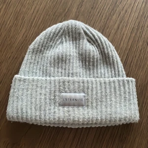Ljusgrå ribbad mössa från H&M - Snygg ljusgrå ribbstickad mössa med uppvikt kant och diskret tygpatch framtill med märket L'ÉTERNITÉ. Perfekt för kyliga dagar och enkel att matcha med olika outfits.