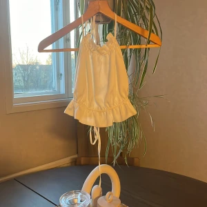 Vit volangprydd linnetopp från Gina Tricot - Supersöt vit linnetopp från Gina Tricot med volangkant nertill och knytband i halsen och ryggen. Toppen är ärmlös och har en lätt glansig finish, perfekt för varma dagar eller sommarens alla tillfällen.