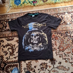 Svart Ed Hardy t-shirt med dödskalletryck - Säljer en svart t-shirt från Ed Hardy med stort tryck av en dödskalle med hatt och smycken på framsidan. T-shirten har korta ärmar och rund hals. Ed Hardy-logga i guld på både fram- och baksidan.