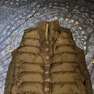 Svart dunväst från Zavetti Canada - Säljer en svart dunväst från Zavetti Canada med hög krage och dragkedja framtill. Västen har broderad logotyp på bröstet och diskreta fickor på sidorna. Perfekt för lager-på-lager och stilren look. Köptes för 850kr och säljs för 650kr, kan gå ner lite i pris om snabb affär. 