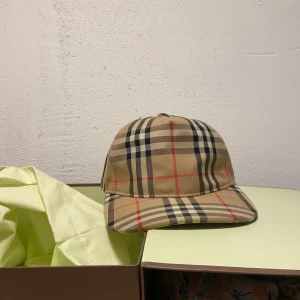 Beige rutig keps från Burberry - Säljer en klassisk beige keps från Burberry med det ikoniska rutmönstret i svart, rött och vitt. Kepsen har böjd skärm och justerbar baksida med svart spänne. Märkeslapp med texten 'Burberry London England' baktill. Box plus påse följer med inget kvitto med den äkta keps pris kan diskuteras 
