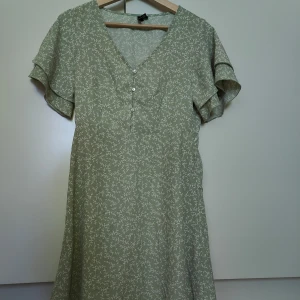 Grön blommig klänning med volangärmar - Söt grön klänning med vitt blommönster och v-ringning. Klänningen har korta volangärmar, knappar framtill och en lätt utställd kjol. 
