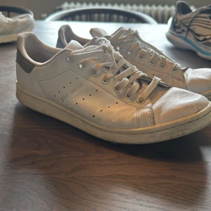 Vita Adidas Stan Smith sneakers - Klassiska vita Adidas Stan Smith sneakers med grå detalj på hälen och perforerade ränder på sidorna. Skorna har snörning och platt sula, perfekt för en stilren och tidlös look.