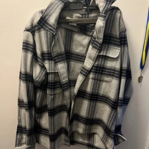 Grå och svart rutig overshirt från Samsoe  - Säljer en snygg overshirt i grått och svart rutigt mönster från Samsoe. Skjortan har långa ärmar, klassisk krage och knappar framtill. Perfekt att slänga över en t-shirt. Använd fåtal gånger. Orginalpris: 1999kr