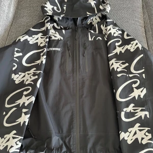 Cortiez Elitework - Säljer en svart vindjacka med vit graffiti-liknande print och huva från Cortiez. Jackan har dragkedja framtill och två bröstfickor med dragkedja. Perfekt för dig som gillar streetwear och vill sticka. All print är även reflex 
