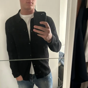 Svart stickad skjorta från COS - Stilren stickad  svart skjorta från COS med klassisk krage och knäppning framtill. Skjortan har lång ärm och är tillverkad i ett mjukt material som passar perfekt till jeans eller chinos.