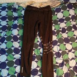 Svarta träningstights från Puma - Svarta tights från Puma med logga längs ena benet. De har en tight passform och är tillverkade i ett stretchigt syntetmaterial, perfekta för träning eller löpning. Reflekterande detalj vid benslutet för extra synlighet.