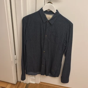 Denim skjorta från Scotch & Soda - Snygg denim skjorta från Scotch & Soda med diskret fiskbensmönster. Klassisk krage, bröstficka och knäppning framtill. Perfekt för dig som gillar stilrena plagg med en twist. Storlek M.