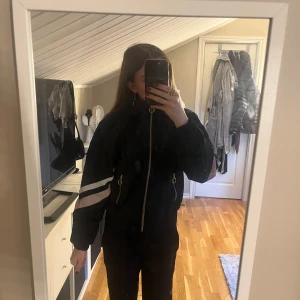 Svart bomberjacka med dragkedja och vita detaljer - Säljer en svart bomberjacka med guldiga dragkedjor och vita samt ljusrosa ränder på båda ärmarna. Jackan har hög krage och resår vid ärmslut och midja. Perfekt för dig som vill ha en jacka som går att sporta i. 